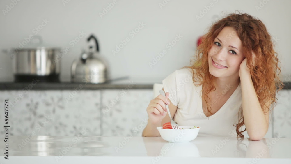 Pretty young woman in kitchen sitting at table with bowl of salad and smiling Pretty young woman in kitchen sitting at table with bowl of salad and smiling