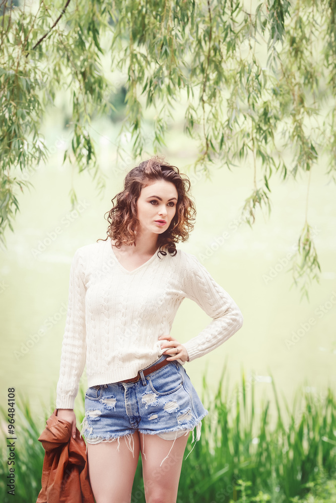 Portrait of beautiful Caucasian young girl woman with wavy curly hair in  white sweater, blue denim shorts and tall leather boots, holding brown  jacket, posing outside in park, summer autumn look Stock
