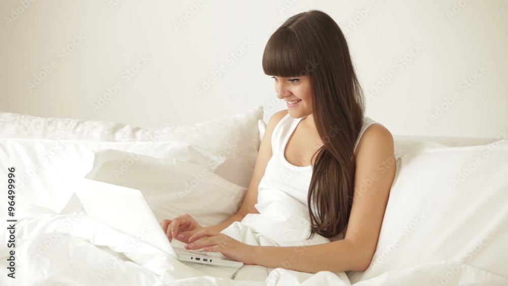 Cheerful girl relaxing in bed using laptop looking at camera and smiling Cheerful girl relaxing in bed using laptop looking at camera and smiling