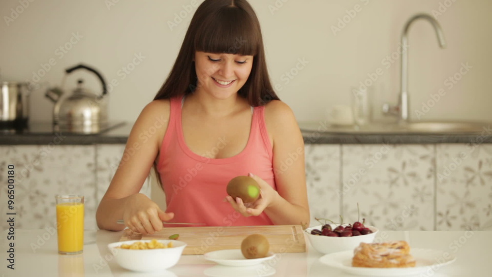 Cheerful girl sitting at kitchen table and peeling kiwi fruit Cheerful girl sitting at kitchen table and peeling kiwi fruit
