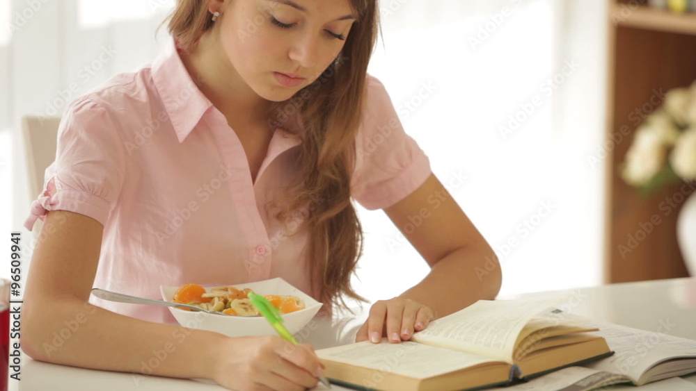 Pretty girl sitting at table writing in workbook looking at camera and smiling. Panning camera