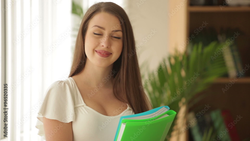 Attractive young woman standing in front of window holding paper folders and smiling at camera Attractive young woman standing in front of window holding paper folders and smiling at camera