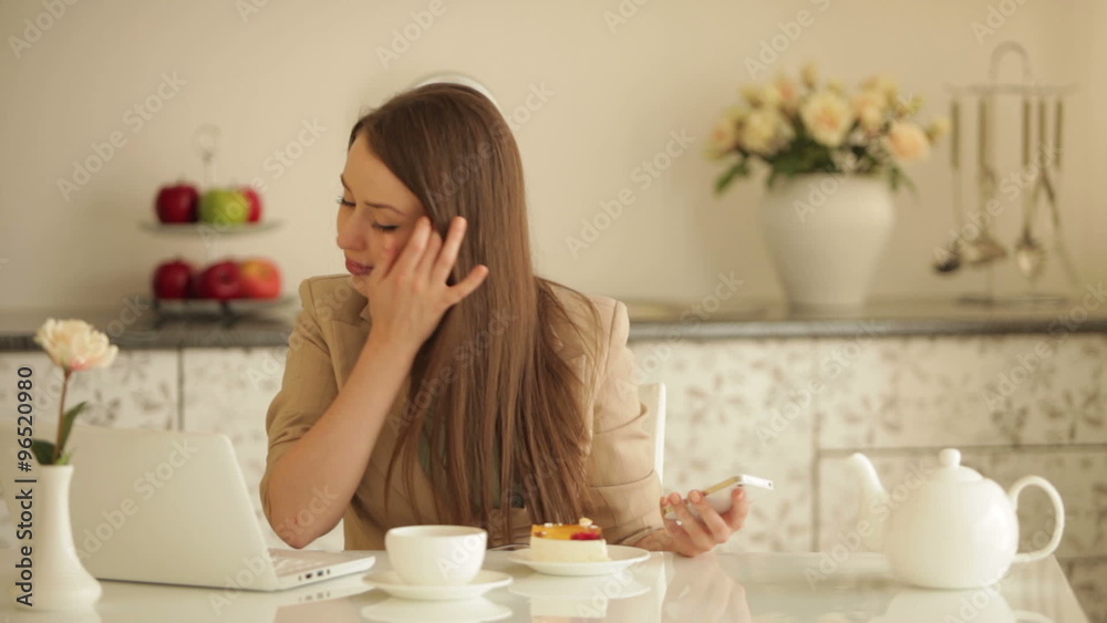 Beautiful girl sitting at kitchen table with laptop holding mobile phone and eating cake Beautiful girl sitting at kitchen table with laptop holding mobile phone and eating cake