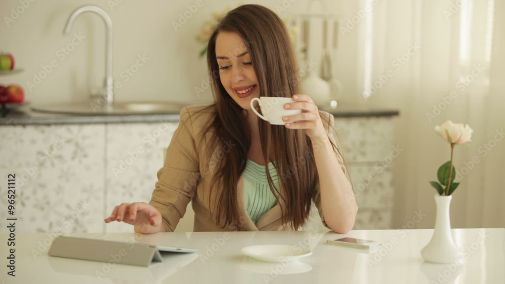 Beautiful girl sitting at table using touchpad drinking tea and smiling at camera Beautiful girl sitting at table using touchpad drinking tea and smiling at camera