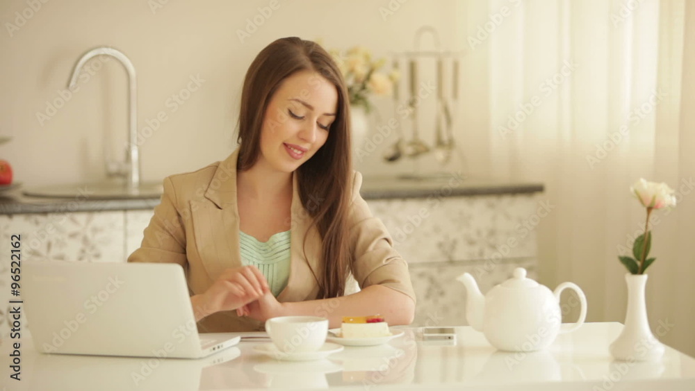 Charming young woman sitting at kitchen table with laptop eating cake drinking tea and smiling Charming young woman sitting at kitchen table with laptop eating cake drinking tea and smiling