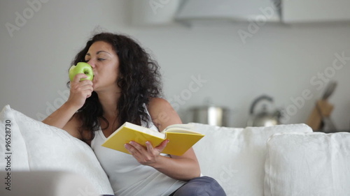 Pretty brunette girl chilling on sofa reading book and eating apple