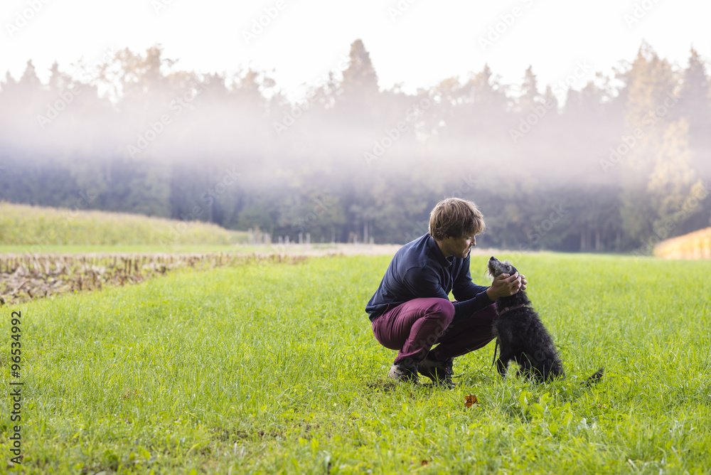 Young man crouching to pet his black dog in a beautiful green me Young man crouching to pet his black dog in a beautiful green me