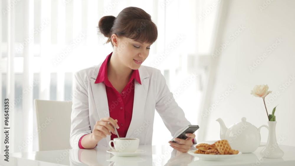 Beautiful young woman sitting at table with cup of tea and using cellphone Beautiful young woman sitting at table with cup of tea and using cellphone
