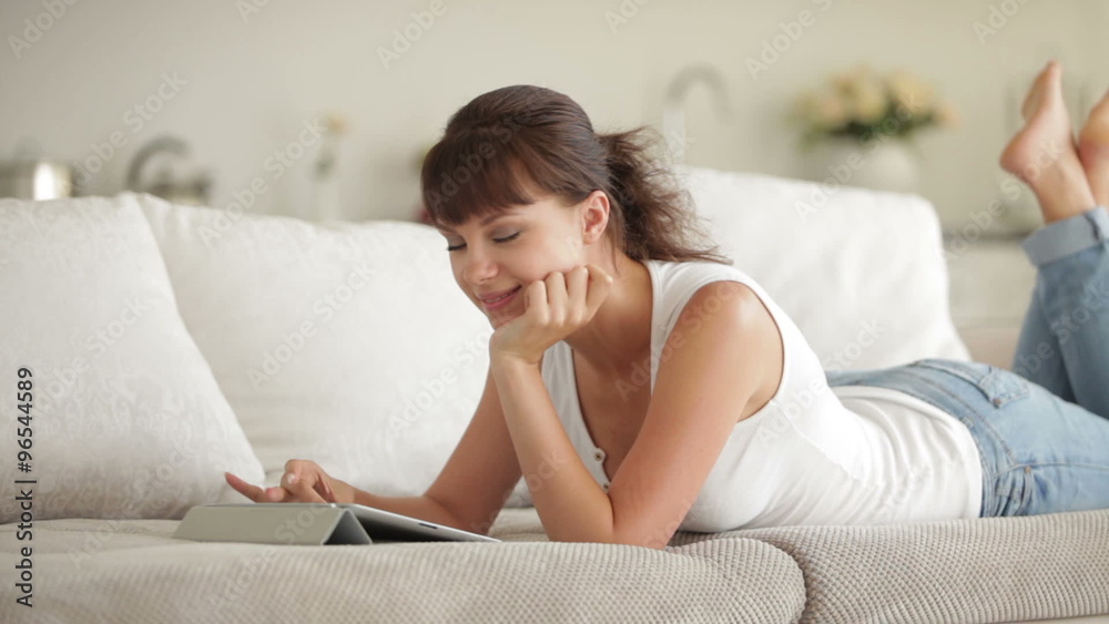 Beautiful girl lying on sofa using touchpad and smiling at camera Beautiful girl lying on sofa using touchpad and smiling at camera