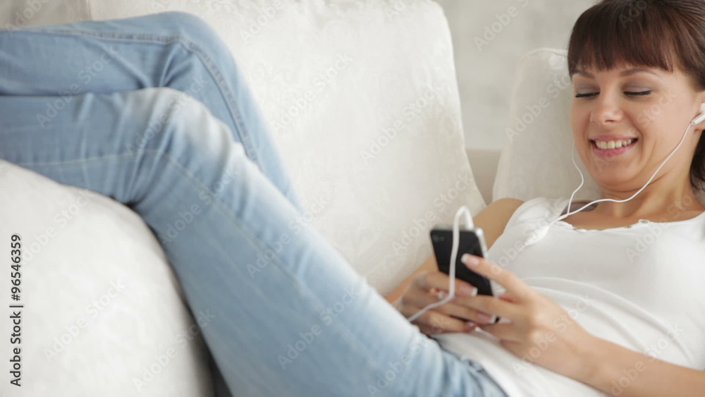 Attractive young woman in headphones sitting on sofa listening to music on mobile phone and smiling Attractive young woman in headphones sitting on sofa listening to music on mobile phone and smiling