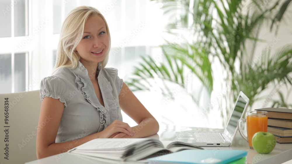 Cute blonde girl sitting at table studying with books and laptop and smiling at camera