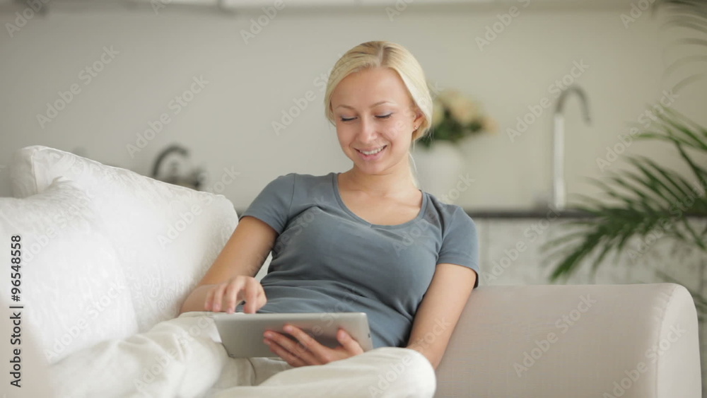 Beautiful young woman relaxing on sofa using touchpad and smiling at camera Beautiful young woman relaxing on sofa using touchpad and smiling at camera