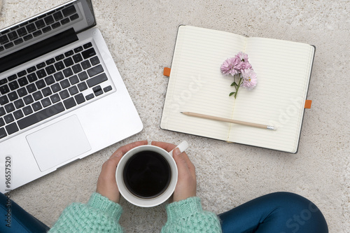 Laptop, notebook and a cup of coffee in girl's hands sitting on white carpet