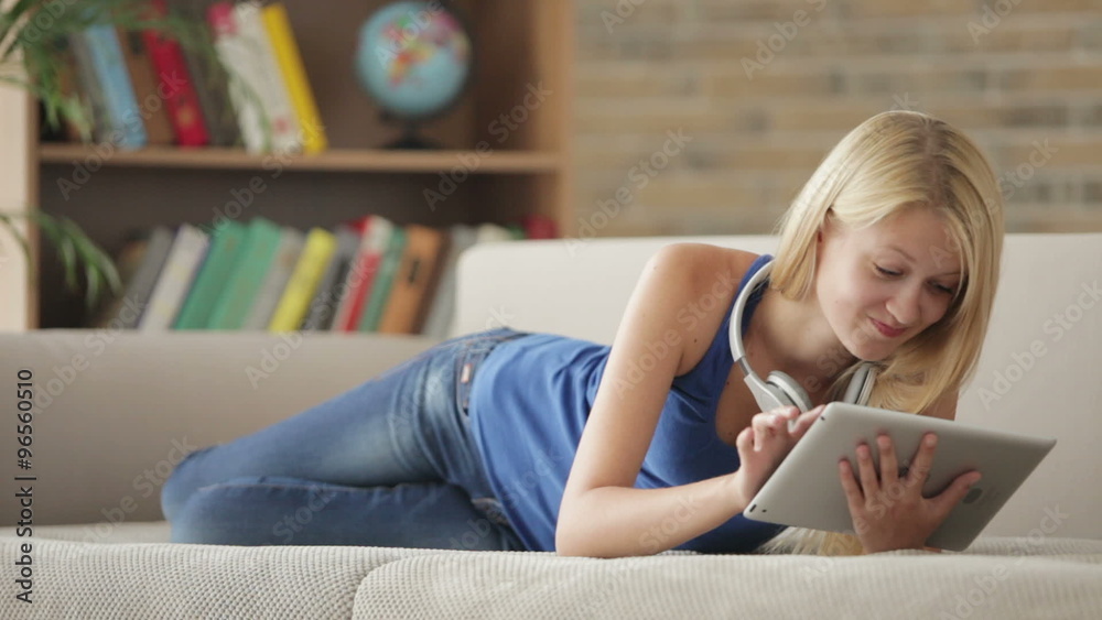 Attractive girl wearing headset lying on couch using touchpad looking at camera and smiling Attractive girl wearing headset lying on couch using touchpad looking at camera and smiling