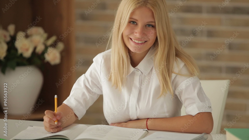 Cheerful girl sitting at table with books studying writing in notebook and smiling at camera. Panning camera