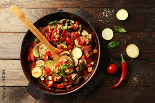 Tasty vegetarian ratatouille made of eggplants, squash, tomatoes and onions in black cast iron pan, on wooden table background photo