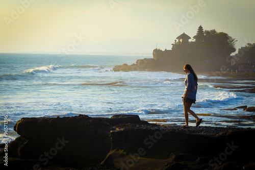 Young woman walking a ocean coastline with the hindu temple Pura Tanah Lot in background, Bali, Indonesia