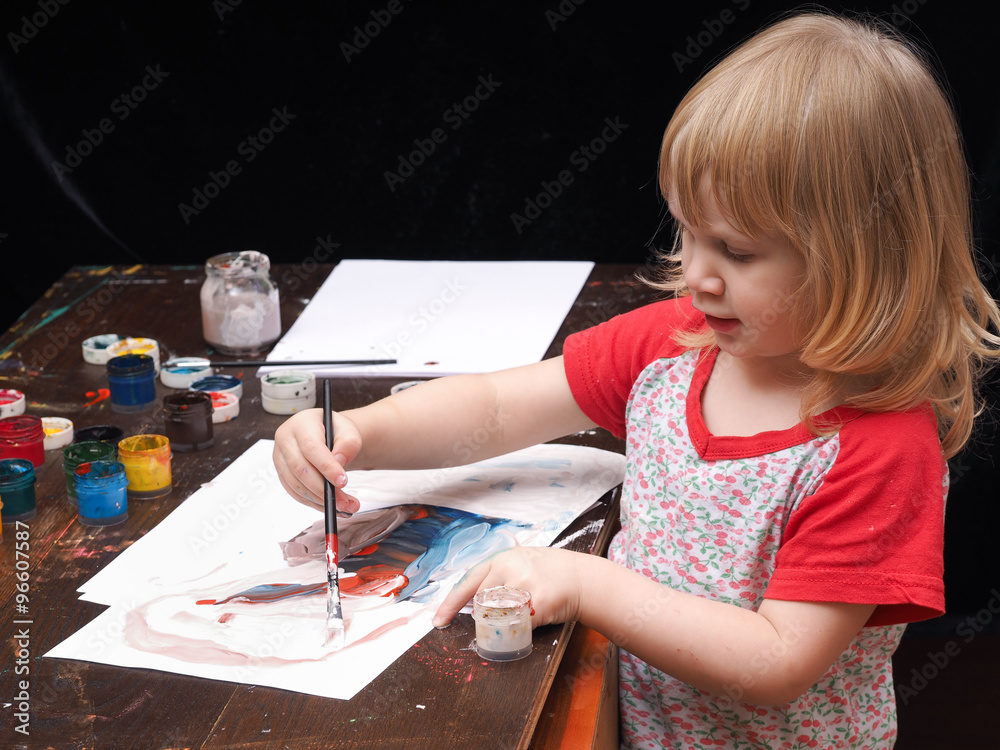 Fotografia do Stock: Маленький ребенок рисует красками, гуашью. Темный стол  в краске, банки с краской. Девочка рисует кистью на белой бумаге. Черный  фон. У девочки светлые волосы, она очень мала | Adobe