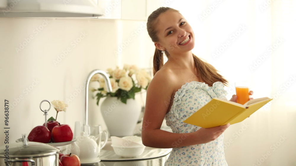 Pretty girl standing in kitchen reading book holding glass of juice and smiling at camera. Panning camera