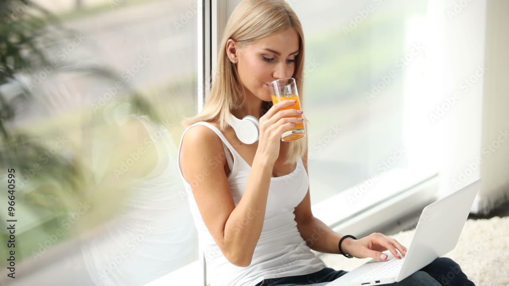 Pretty girl sitting by window using laptop drinking juice and smiling Pretty girl sitting by window using laptop drinking juice and smiling