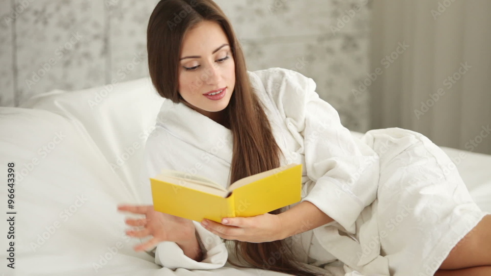Pretty girl in bathrobe lying in bed reading book looking at camera and smiling. Panning camera Pretty girl in bathrobe lying in bed reading book looking at camera and smiling. Panning camera