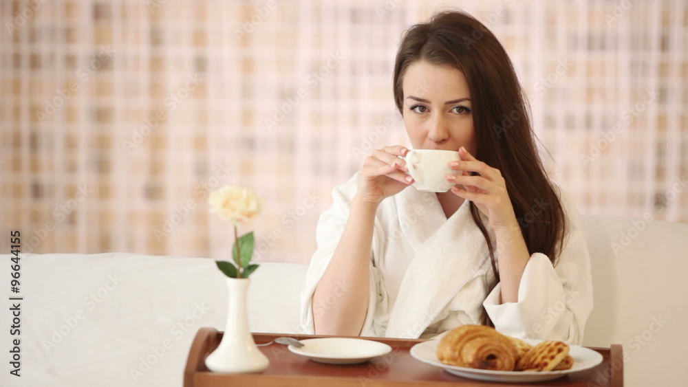 Cute girl in bathrobe sitting in bed drinking tea looking at camera and smiling. Panning camera Cute girl in bathrobe sitting in bed drinking tea looking at camera and smiling. Panning camera