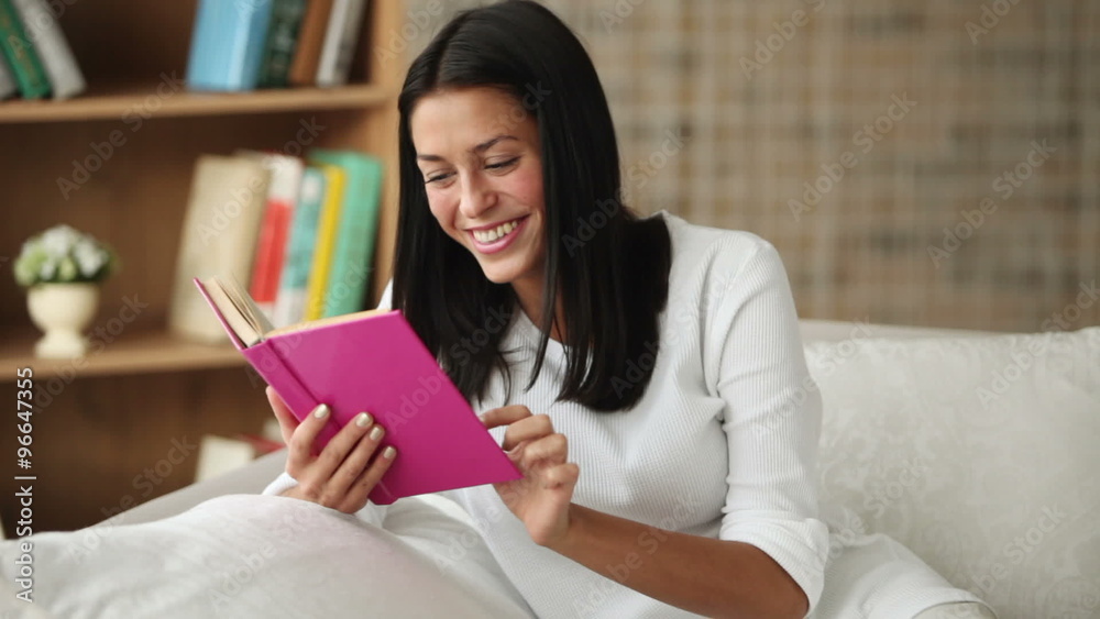 Charming girl sitting on sofa reading book closing it looking at camera and smiling. Panning camera Charming girl sitting on sofa reading book closing it looking at camera and smiling. Panning camera