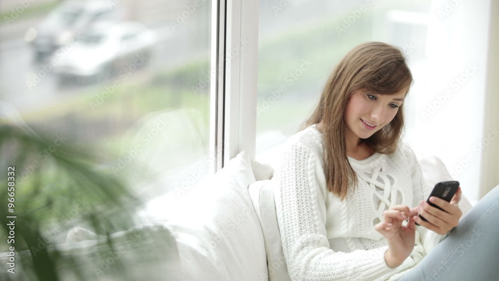 Pretty girl sitting by window using mobile phone looking at camera and smiling Pretty girl sitting by window using mobile phone looking at camera and smiling