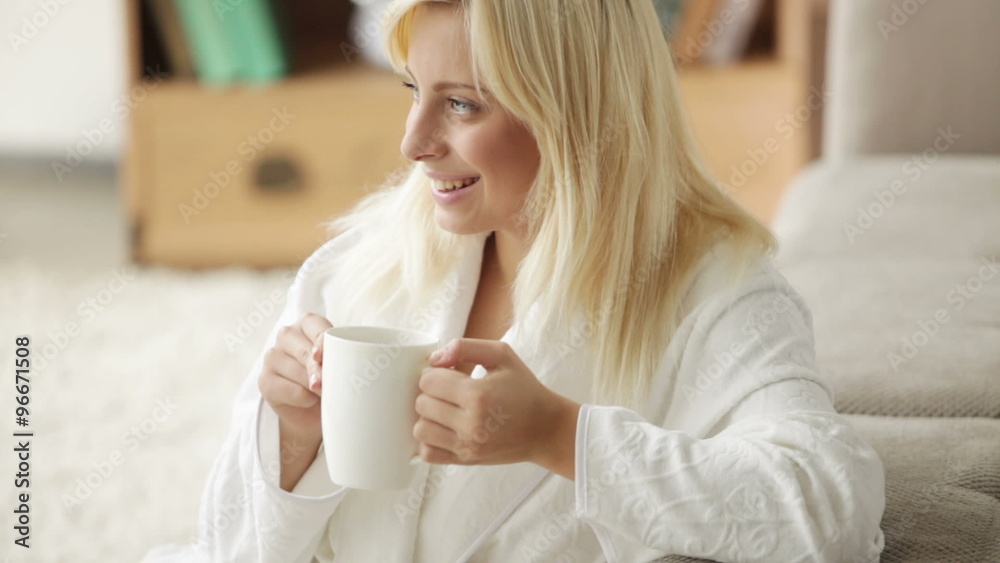 Cheerful young woman wearing bathrobe sitting on floor drinking from cup looking at camera and smiling. Panning camera Cheerful young woman wearing bathrobe sitting on floor drinking from cup looking at camera and smiling. Panning camera