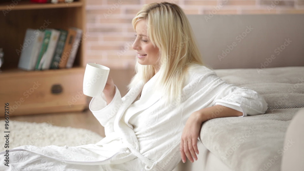 Cute young woman in bathrobe sitting on floor drinking from cup looking at camera and smiling. Panning camera Cute young woman in bathrobe sitting on floor drinking from cup looking at camera and smiling. Panning camera