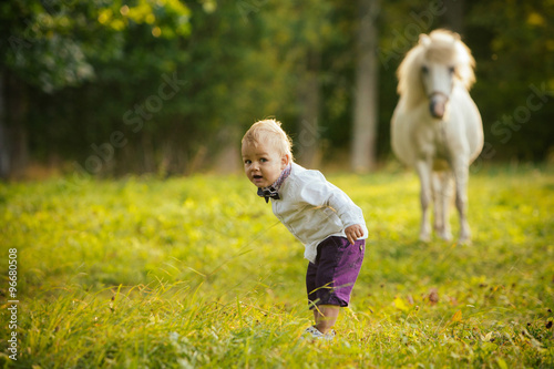 Little boy dressed up in elegant clothes standing in meadow, white pony in the background.