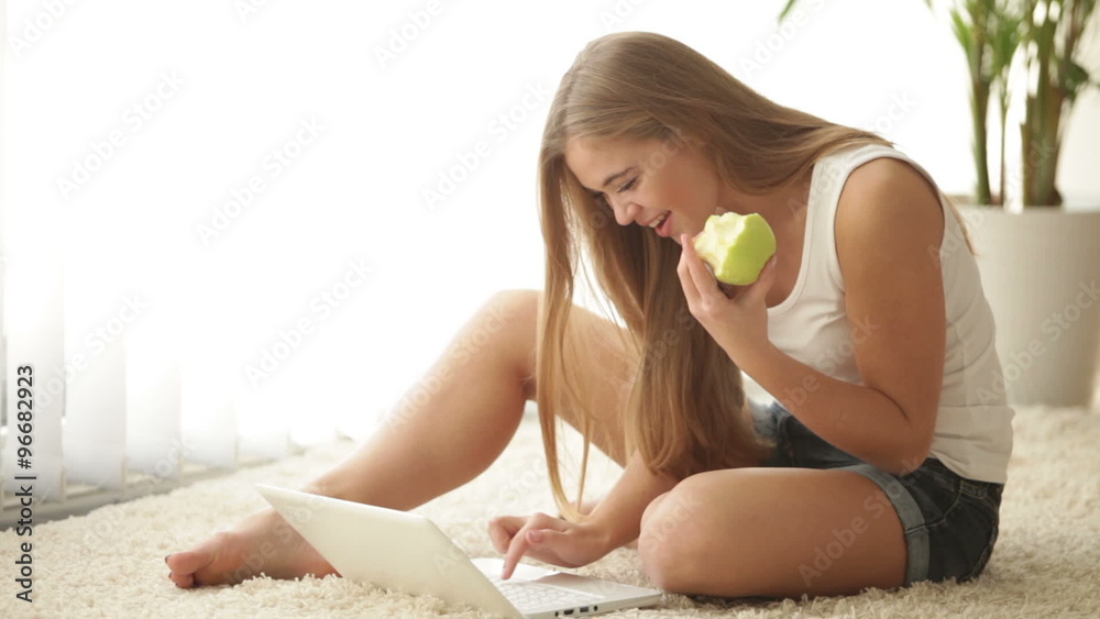 Attractive girl sitting on floor using laptop eating apple and smiling. Panning camera
