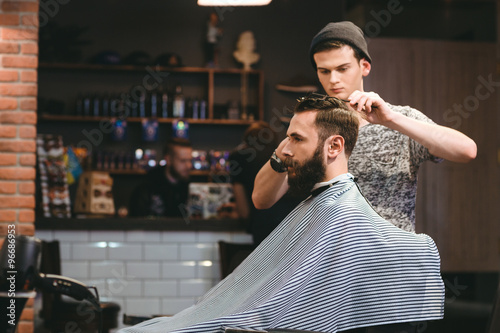 Young barber making haircut of bearded man in barbershop