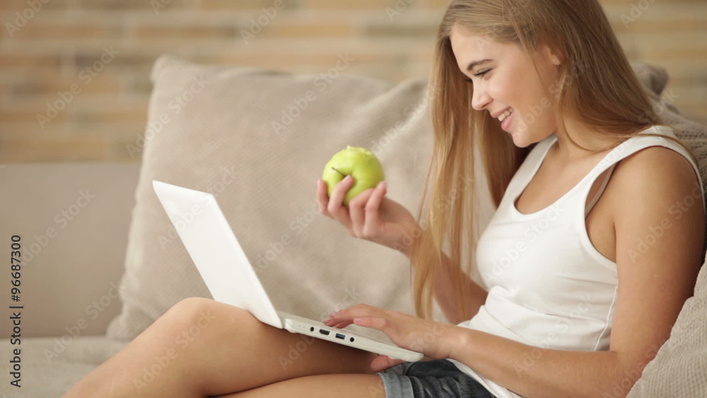 Pretty young woman sitting on sofa using laptop eating apple and smiling. Panning camera