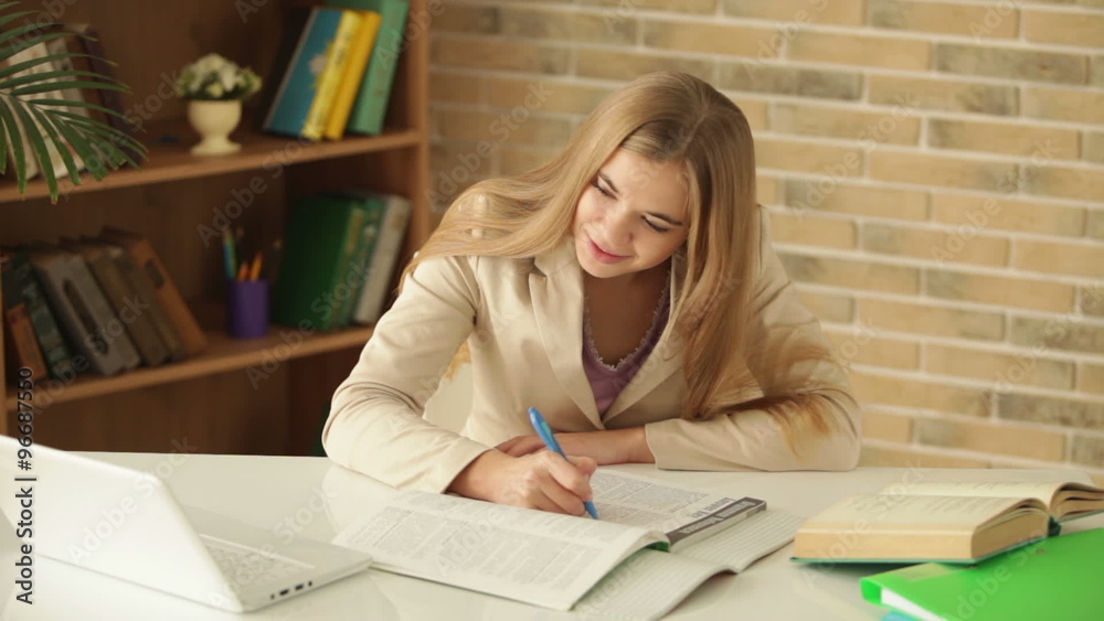 Student girl sitting at table writing in workbook using laptop smiling and showing thumb up at camera Student girl sitting at table writing in workbook using laptop smiling and showing thumb up at camera