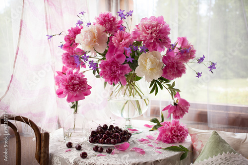   A bouquet of pink peonies, bells in a vase and cherries in a plate on the table by the window. Still life in a country house. 