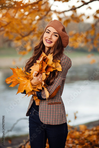 slender young brunette beautiful girl with long hair in a beret with a scarf and coat walks in autumn park with a lot of yellow and orange leaves