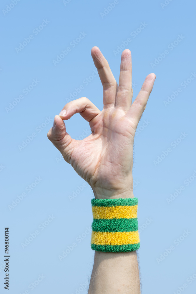 Hand making OK sign with finger and thumb against blue sky wearing a yellow and green sport wristband Hand making OK sign with finger and thumb against blue sky wearing a yellow and green sport wristband