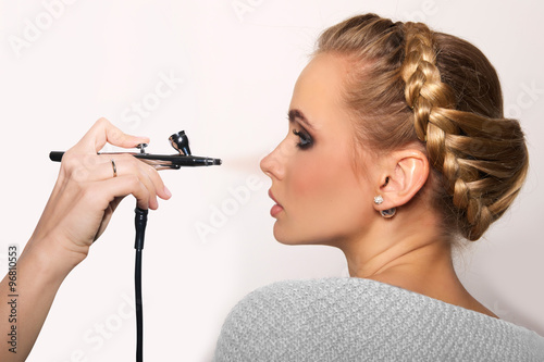 portrait of a beautiful young blonde woman on a light background. there is hand with aerograph making an airbrush make up. hair tied in a braid. copy space..