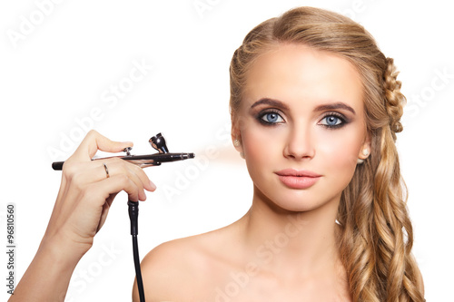 portrait of a beautiful young blonde woman on a light background. there is hand with aerograph making an airbrush make up. hair tied in a braid. copy space..