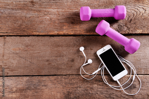 White cellphone with headphones and pink dumb bells on  wooden background