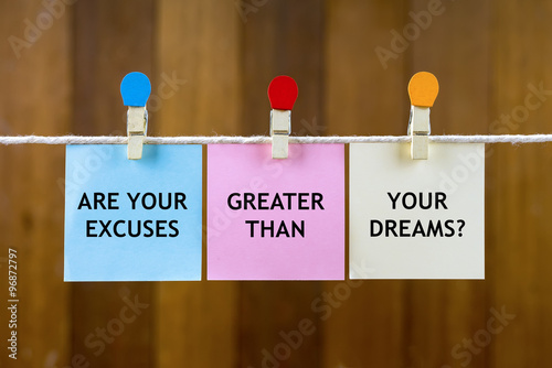 Word quotes of ARE YOUR EXCUSES GREATER THAN YOUR DREAMS? on colorful sticky papers hanging by a rope against blurred wooden background.