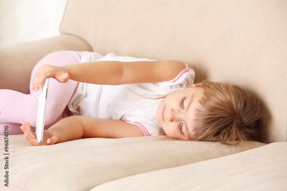 Naklejka premium Beautiful little girl with smart phone lying on sofa, on home interior background