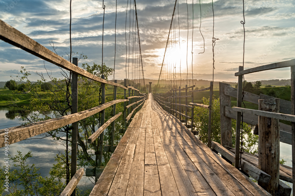 Obraz premium Old lengthy hanging wooden footbridge with rails over river against sunset background. Arkhangelsky region, Russia.