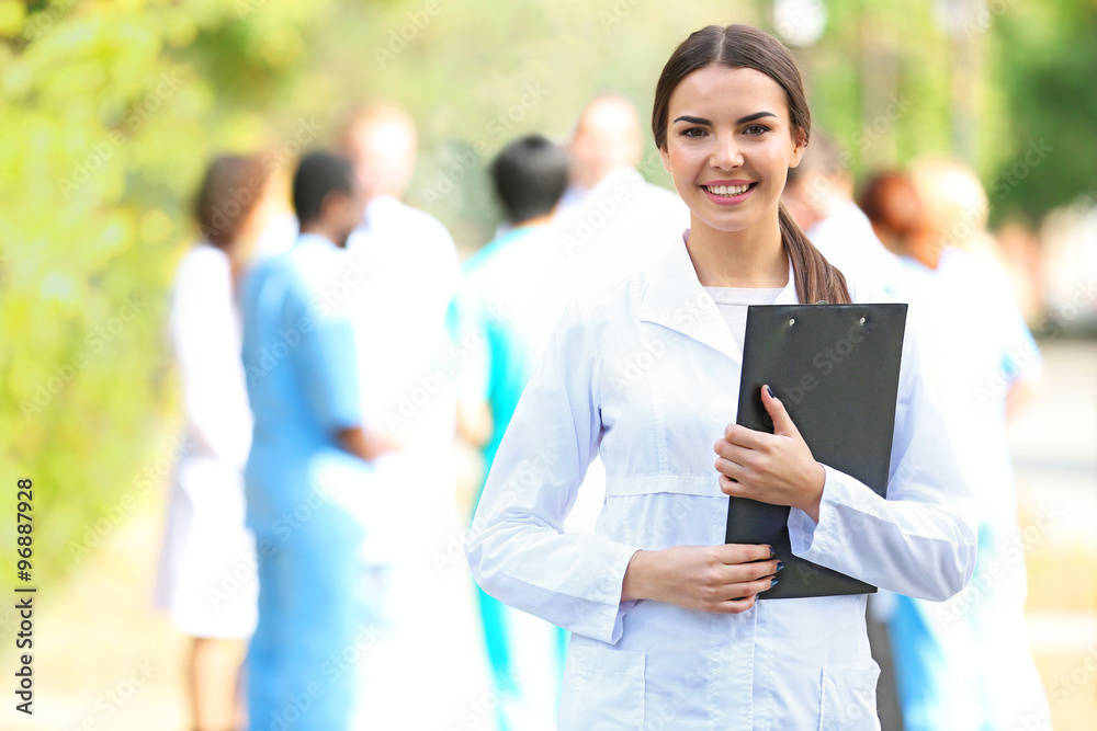 Beautiful young woman doctor with clipboard in hands against group of medics Beautiful young woman doctor with clipboard in hands against group of medics