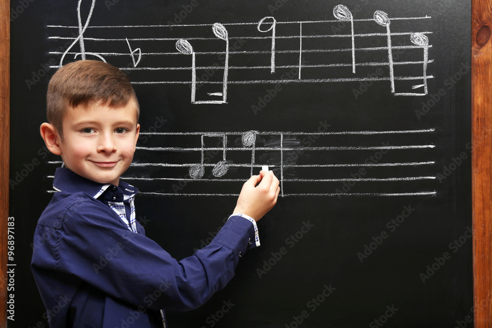 Cute boy writing at the blackboard with musical notes, in the classroom