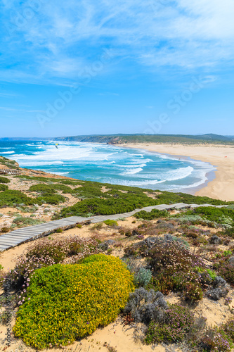 Yellow flowers and wooden walkway to Praia do Bordeira beach with beautiful blue sea view, Algarve region, Portugal