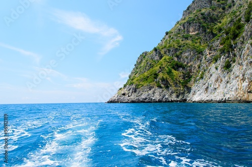 Motorboat trails on a blue sea with steep verdant cliff in Cilento