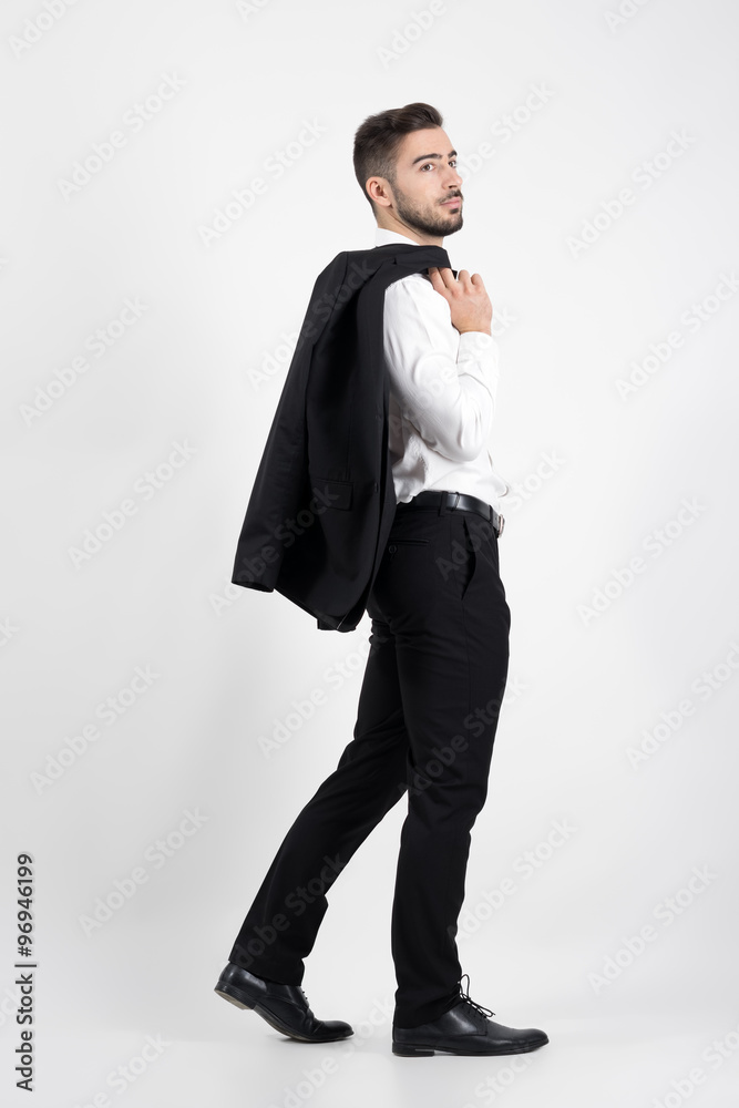 Young elegant luxurious man walking carrying tuxedo over his shoulder looking away. Full body length portrait over gray studio background. Young elegant luxurious man walking carrying tuxedo over his shoulder looking away. Full body length portrait over gray studio background.