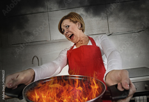 young inexperienced home cook woman in panic with apron holding pot burning in flames with in panic